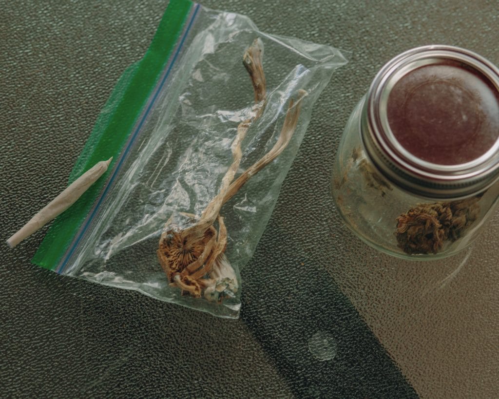 DoubleBlind: Jar of weed, bag of mushrooms and a joint on a table. In this article, Doubleblind explores what you need to know about smoking weed while tripping.