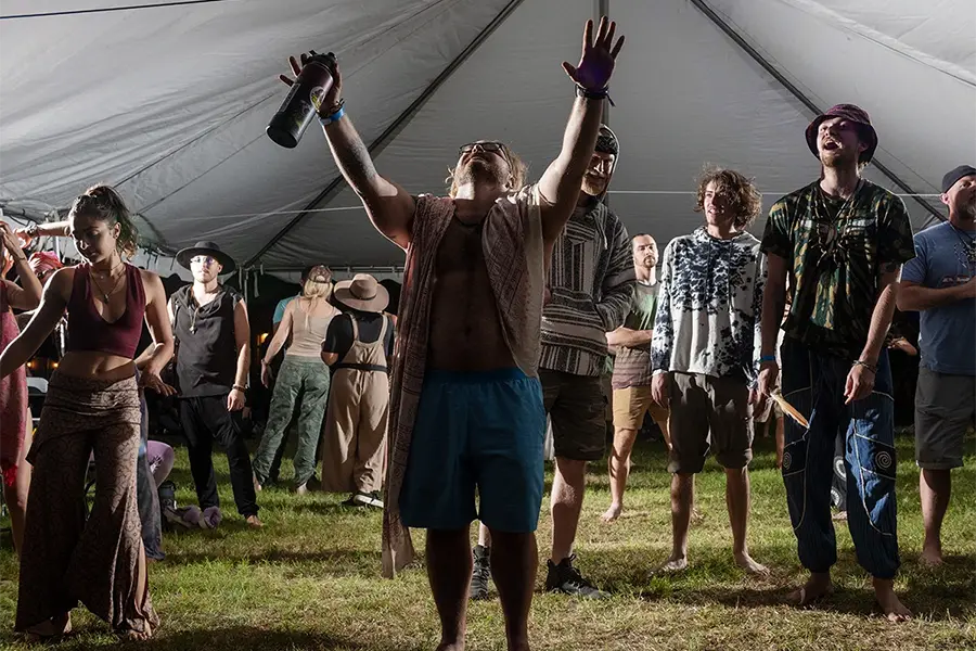 a supporter of the church of psilomethoxin holds both arms up toward the sky in a tent, holding water bottle